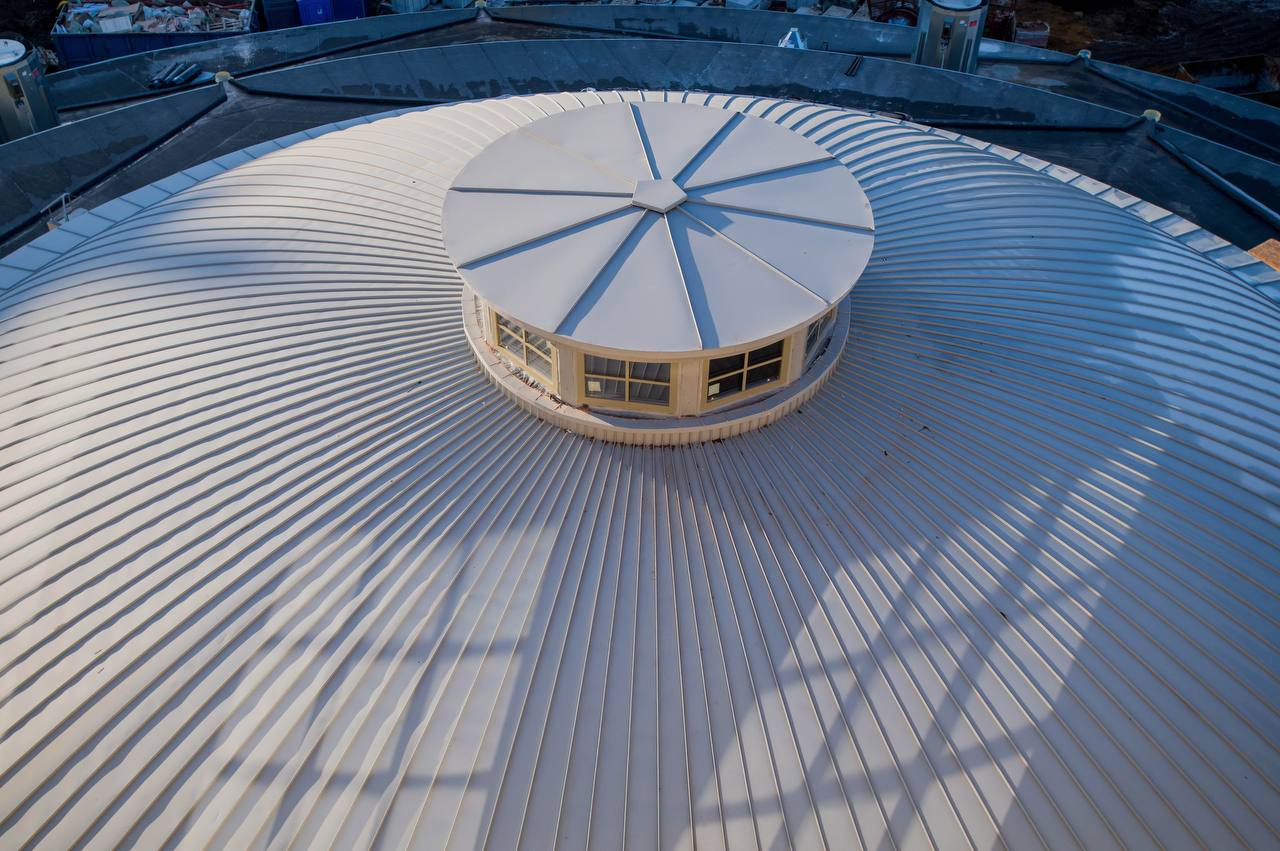 Vue aérienne d'un dôme couvert de bandes métalliques radiales. Au centre, un lanterneau circulaire vitré à couvercle segmenté, entouré d'un toit plat sombre.