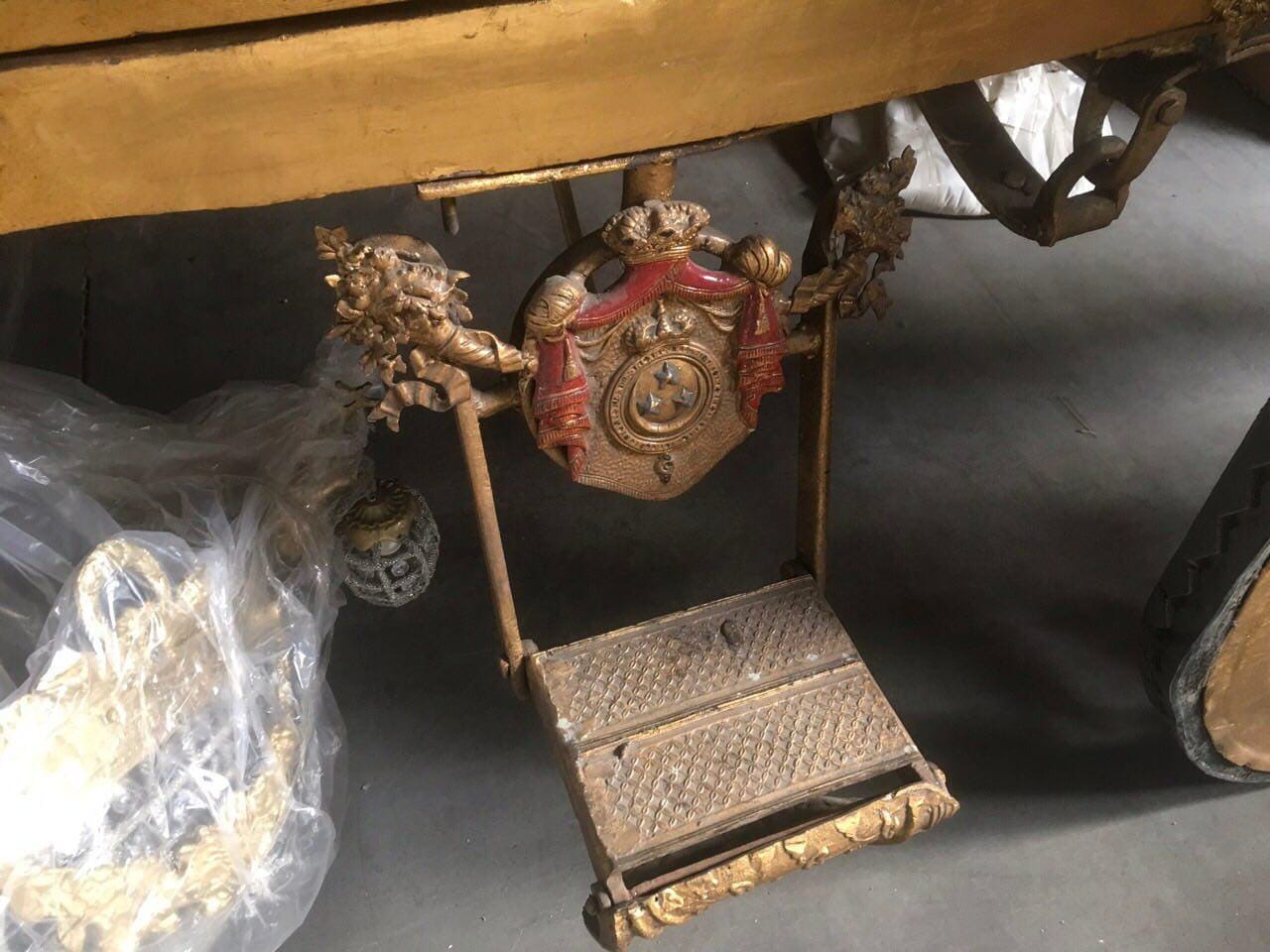 Marchepied rabattable d'un carrosse doré avec un blason en relief : couronne, draperie rouge, trois étoiles sur l'écu. Des éléments de décor sous plastique sont posés à côté.