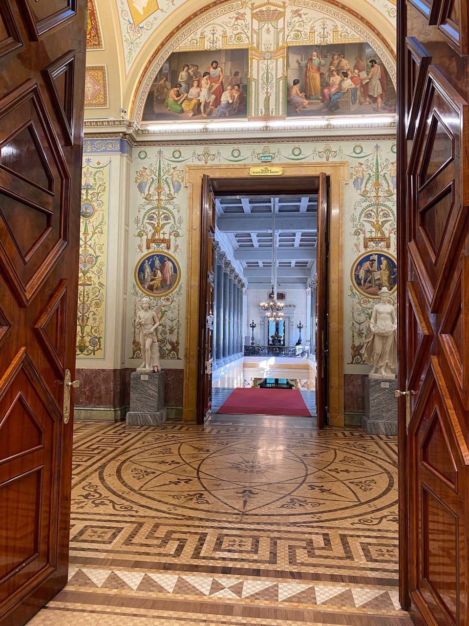 Des portes en bois ouvertes donnent sur une salle aux murs peints, au sol en mosaïque et aux statues de marbre ; au fond, des colonnes grises et un tapis rouge.