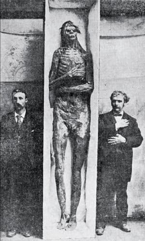 Black-and-white photograph of a mummy standing upright in a niche, flanked by two men in dark suits.