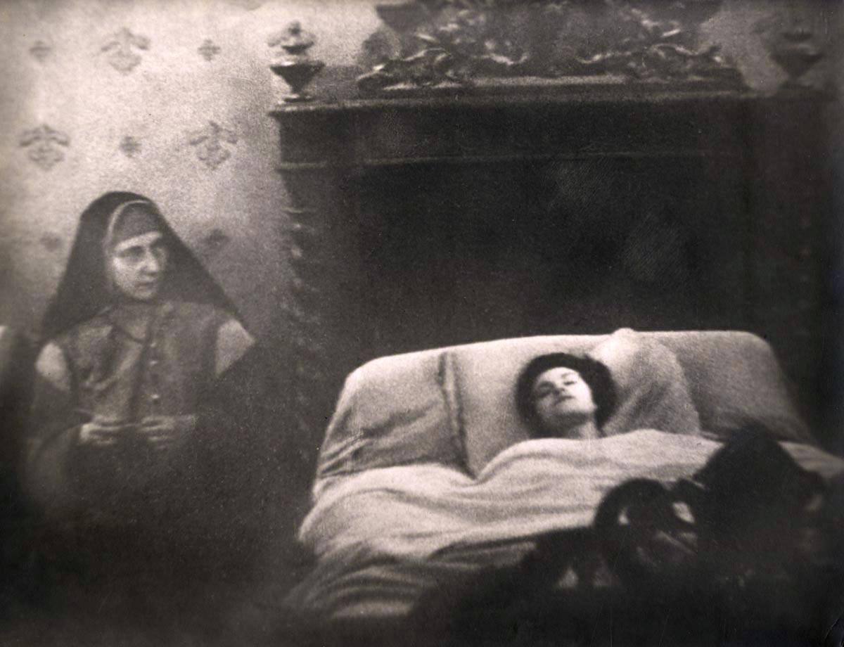 Photo ancienne : une religieuse voilée de noir est assise à gauche ; à droite, une femme brune repose sur des oreillers, les yeux clos. Au fond, meuble sculpté et papier peint fleuri.