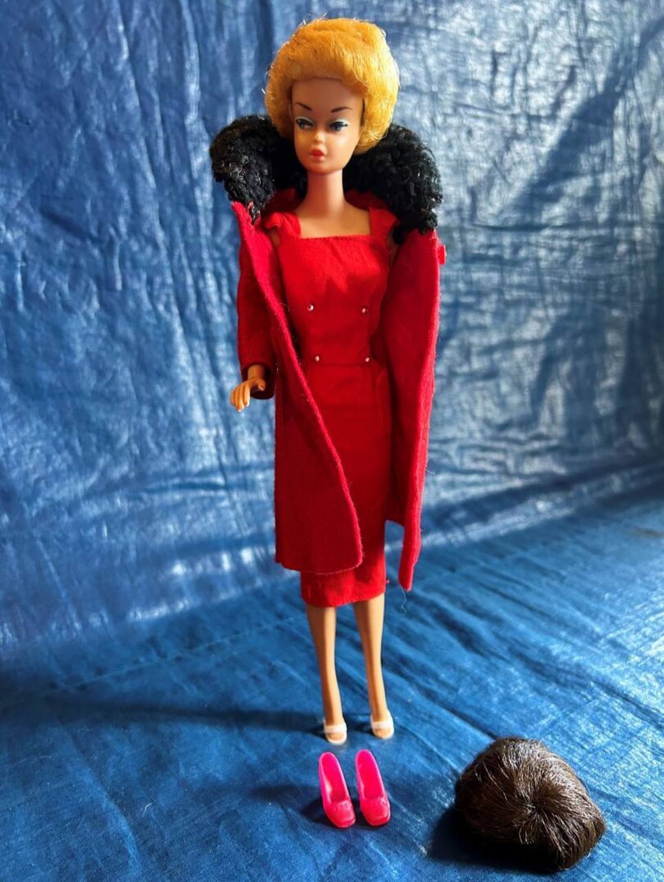 Une poupée blonde en robe rouge et manteau à col noir duveteux se tient sur un tissu bleu. Devant elle : des chaussures roses et une perruque brune.
