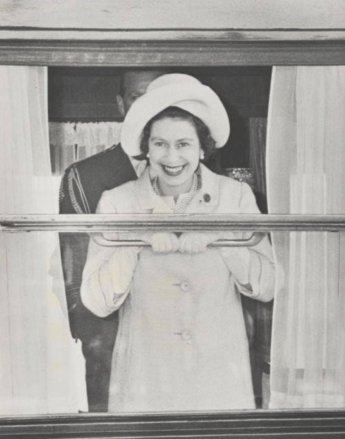 A smiling woman in a light coat and hat holds the rail of a train window, with a man in dark clothing visible behind her.