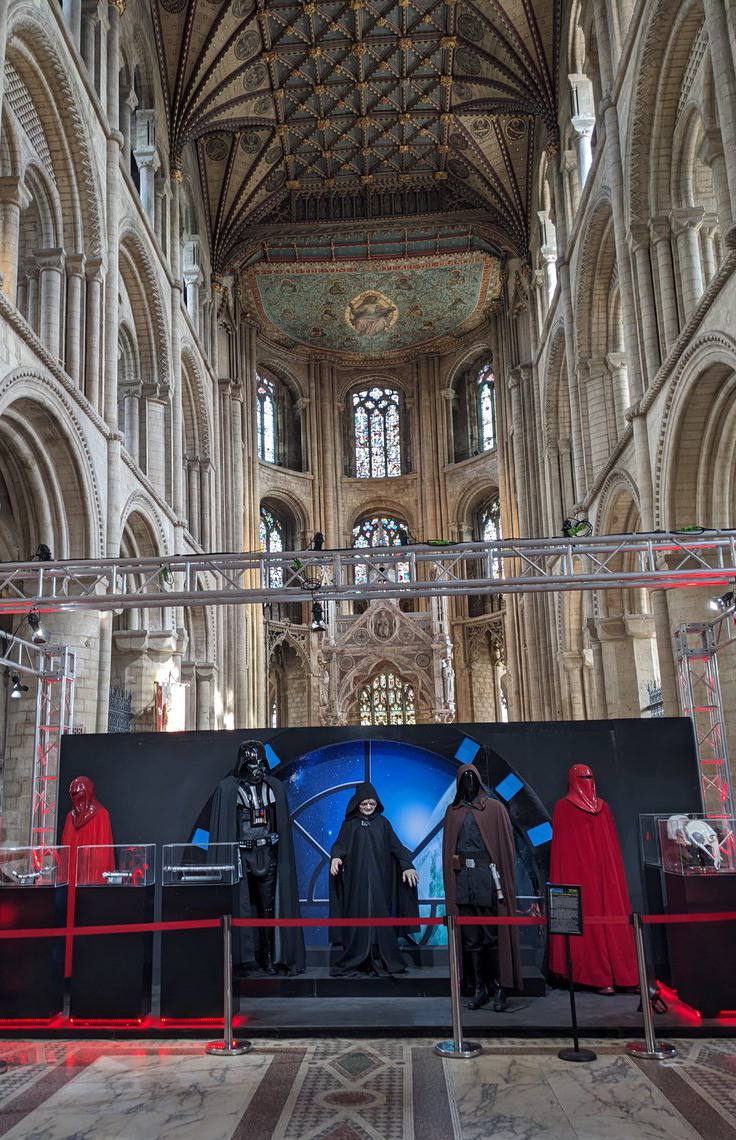 Mannequins en costumes de Dark Vador, de l'Empereur et de gardes rouges derrière un cordon, sous le plafond peint et les vitraux d'une cathédrale.