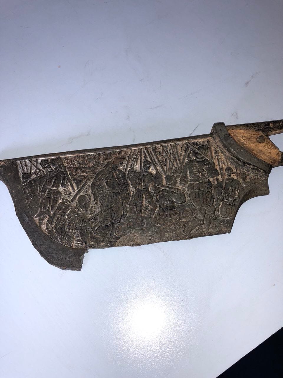 A darkened metal object with a relief scene of soldiers and a wooden part at one edge lies on a light surface.