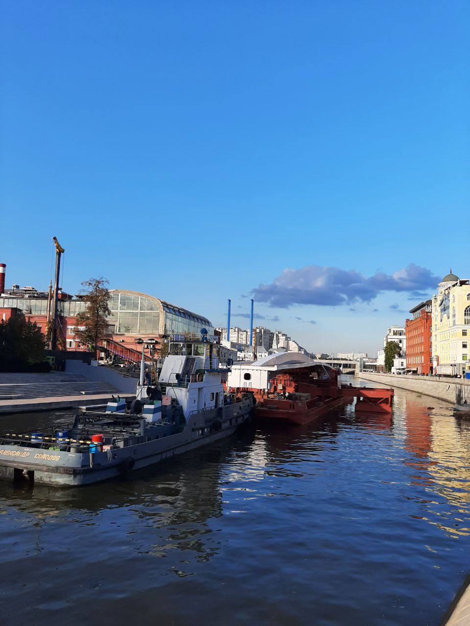 Un remorqueur gris et une barge rouge amarrés près du quai d'un canal. Des bâtiments en brique rouge et une verrière bordent les rives sous un ciel bleu.