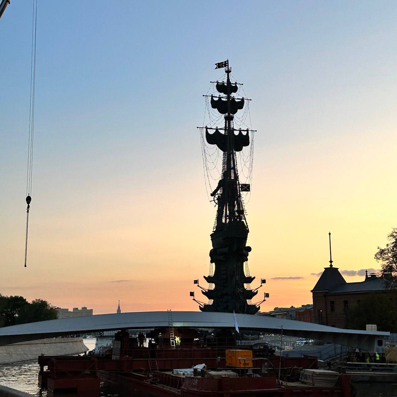 Silhouette sombre d'un monument en forme de navire à voiles sur un ciel orange et bleu ; au premier plan, une travée d'acier sur des barges et un crochet de grue.
