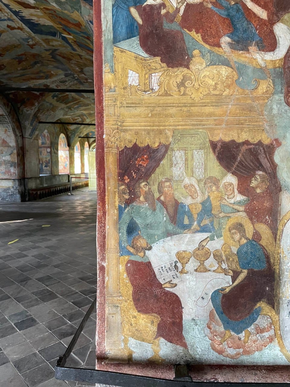 Fresque montrant un groupe attablé devant des coupes, une colonne au premier plan. À gauche, une galerie voûtée aux murs et au plafond peints, au sol dallé.