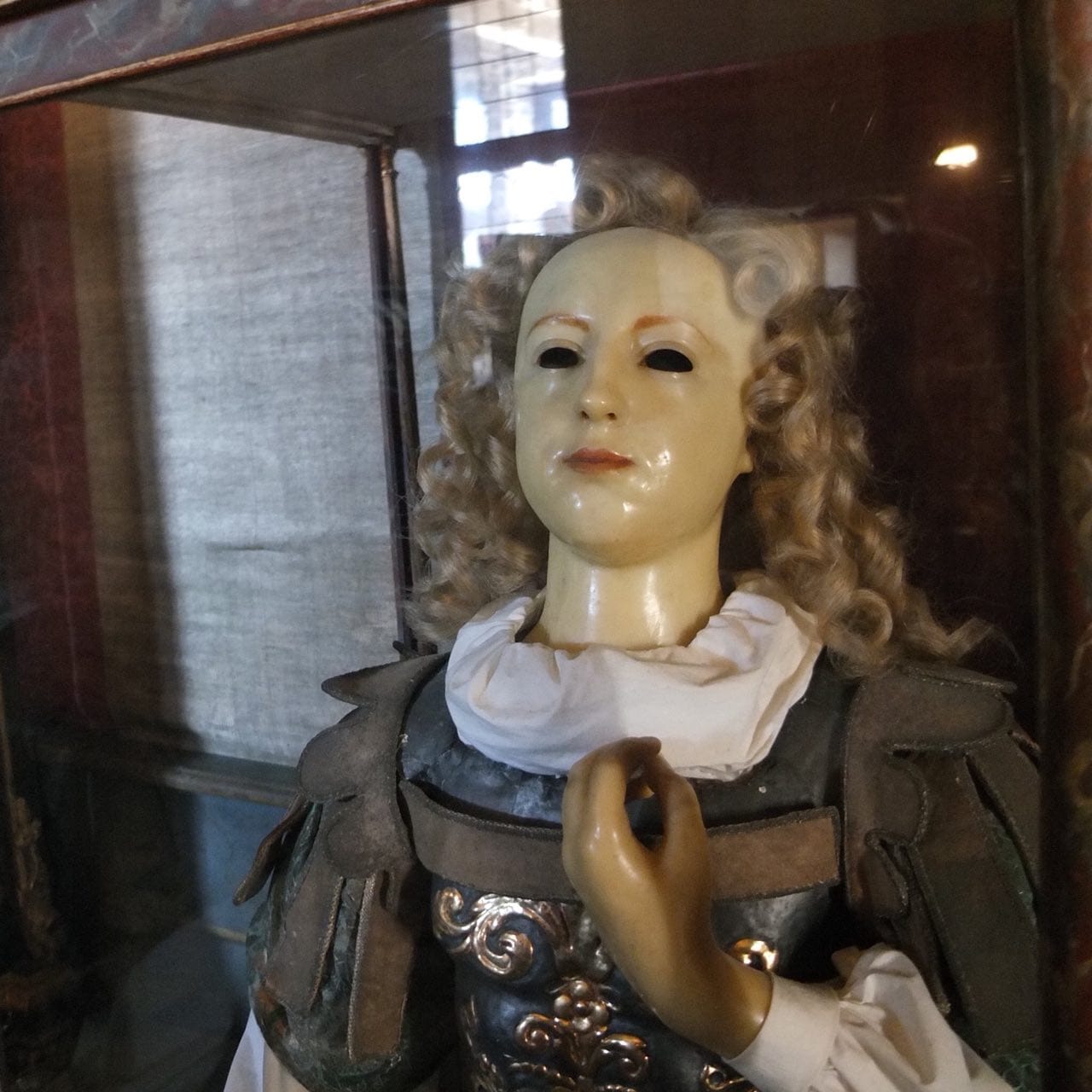 Figure de cire aux orbites vides, aux boucles claires, au col blanc et en armure, derrière la vitre d'une vitrine.