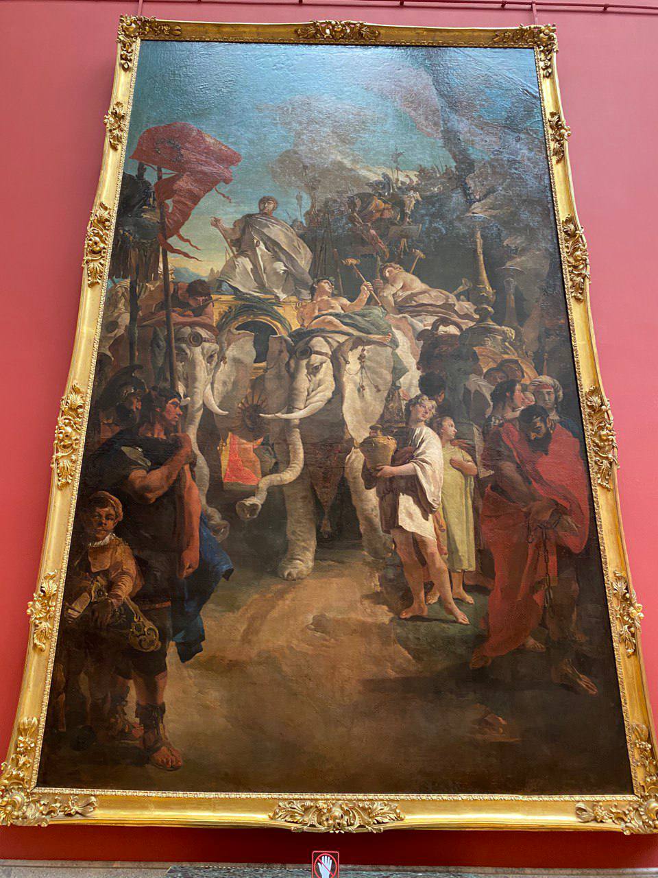Immense toile au cadre doré sur un mur rouge de musée : un homme en toge sur un char tiré par des éléphants blancs, avec soldats, captif enchaîné et jeune homme à la cruche.