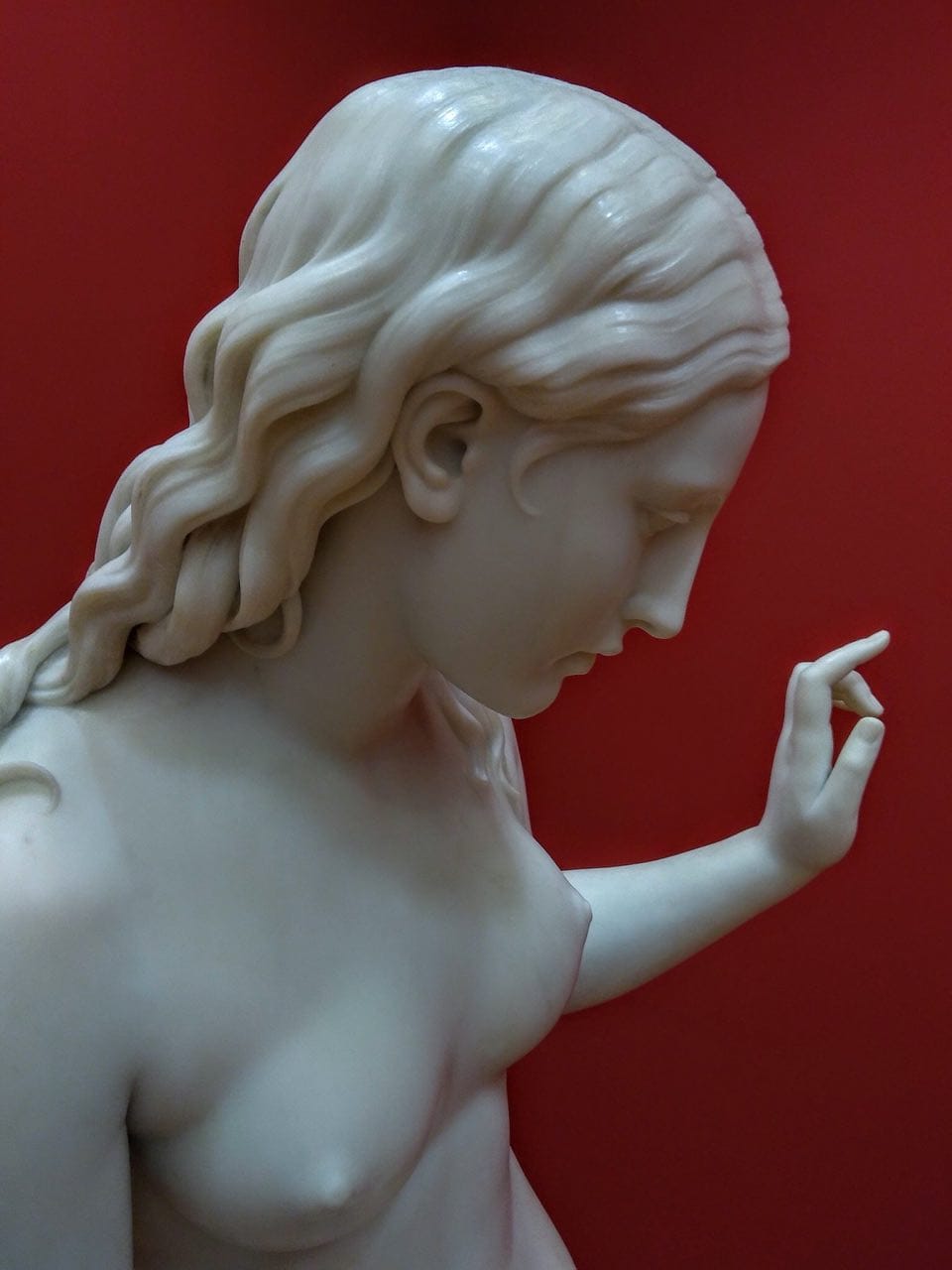 Figure en marbre blanc d'une jeune femme aux cheveux ondulés et dénoués, la tête penchée, une main levée aux doigts à demi repliés. Le fond est rouge vif.