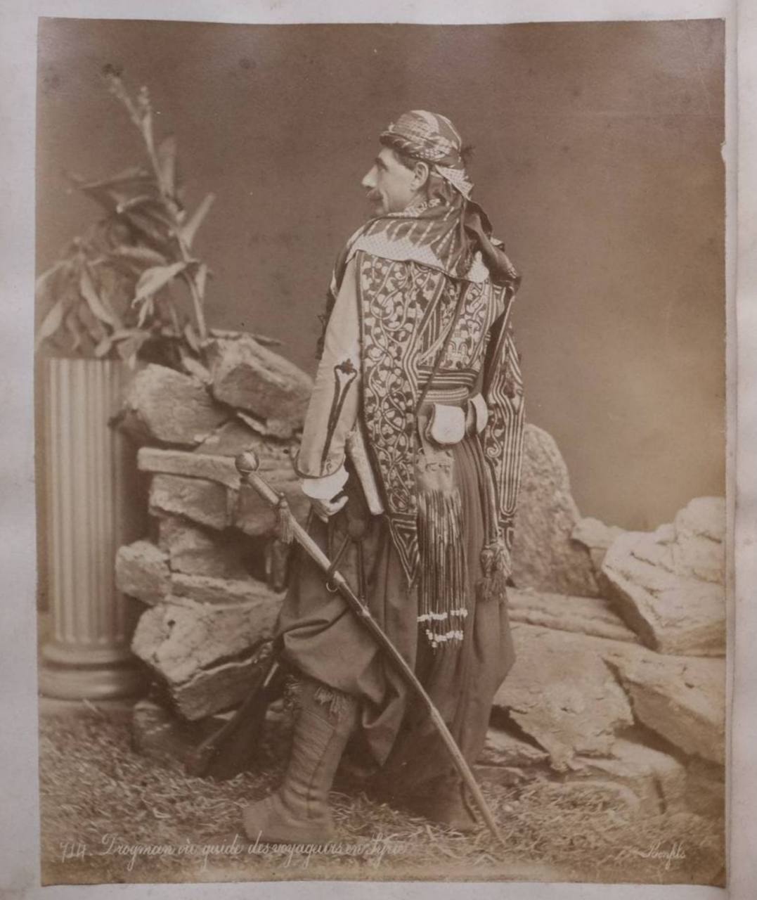 Ancienne photographie d'un homme de profil et de dos, en turban, gilet brodé et sarouel, portant un sabre, devant un décor de studio avec colonne et rochers.