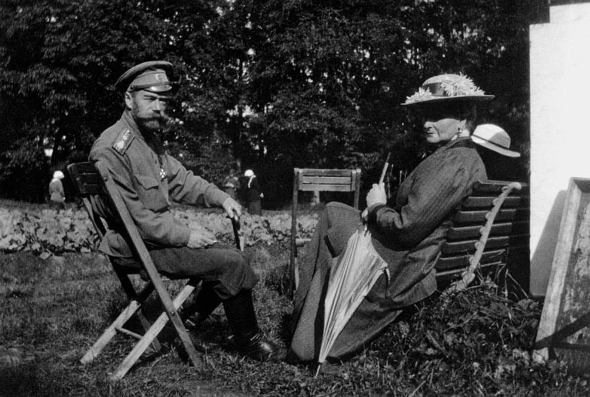 Photographie en noir et blanc d'un homme en uniforme militaire et d'une femme coiffée d'un chapeau, une ombrelle à la main, assis sur des bancs dans un parc.