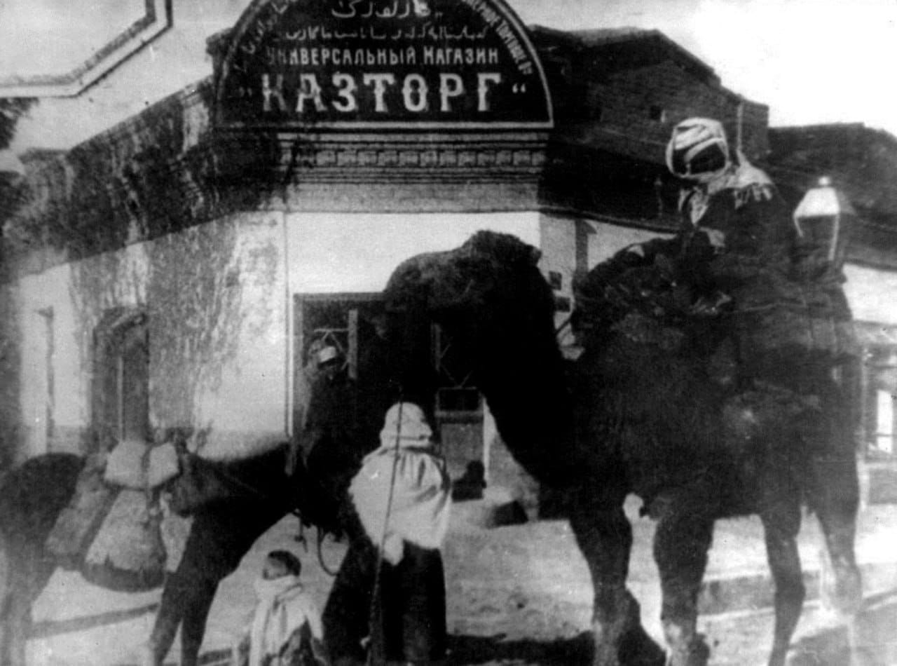 Ancienne photographie d'une enseigne « Magasin universel Kaztorg », avec un chameau monté et des personnes en vêtements chauds devant l'entrée.