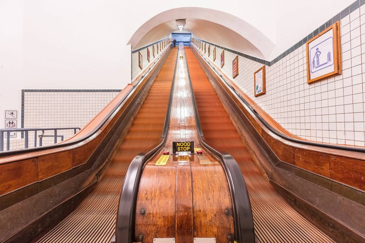 Deux longs escalators en bois montent dans un tunnel carrelé de blanc, séparés par un panneau central en bois portant l'inscription « NOOD STOP ».