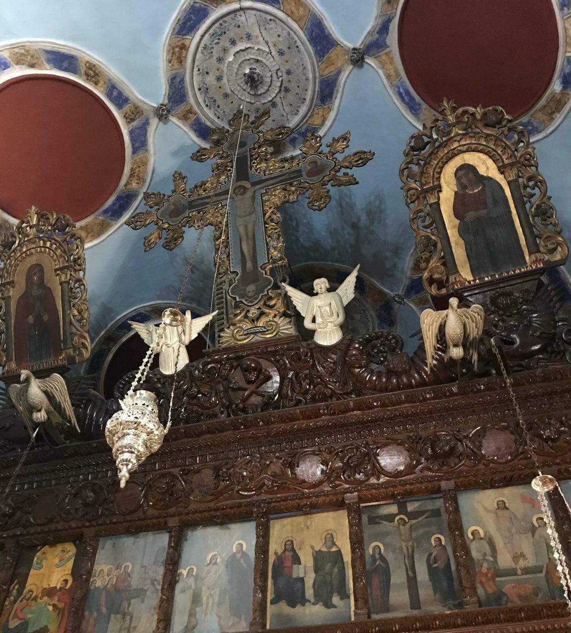 Резной деревянный иконостас с позолоченным крестом с распятием, иконами, фигурами ангелов и птиц на фоне расписного голубого потолка.