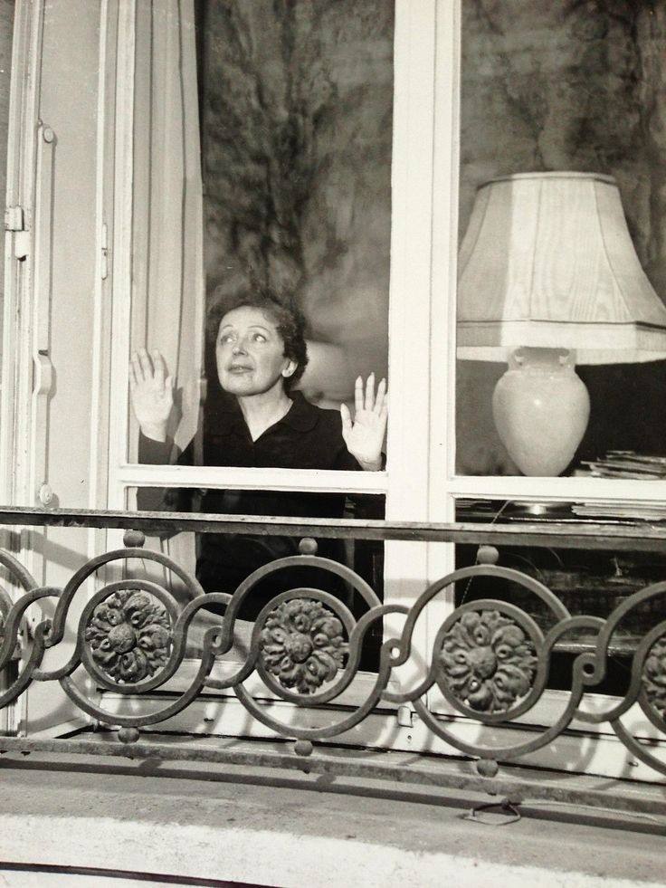Чёрно-белая фотография: женщина в тёмной одежде стоит за окном, подняв ладони к стеклу, перед окном кованая балконная решётка.