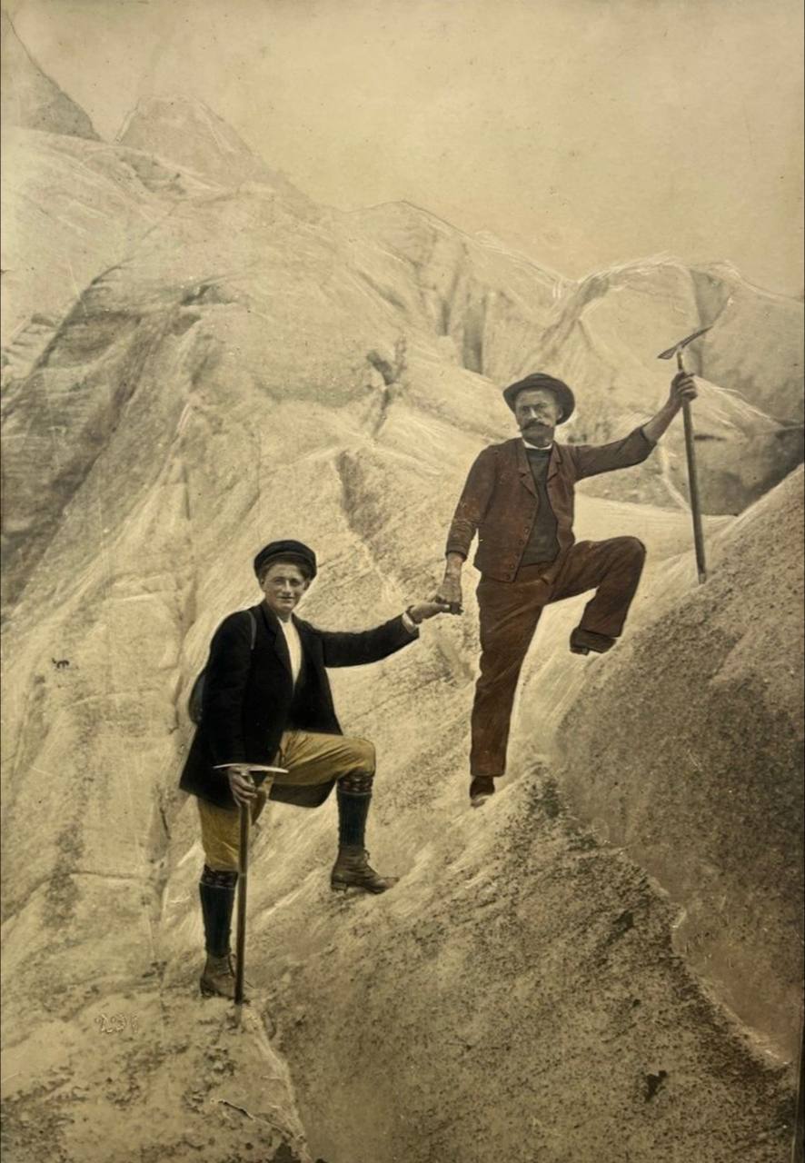 Старая тонированная фотография: двое альпинистов с ледорубами на ледниковом склоне, стоящий выше протягивает руку нижнему.
