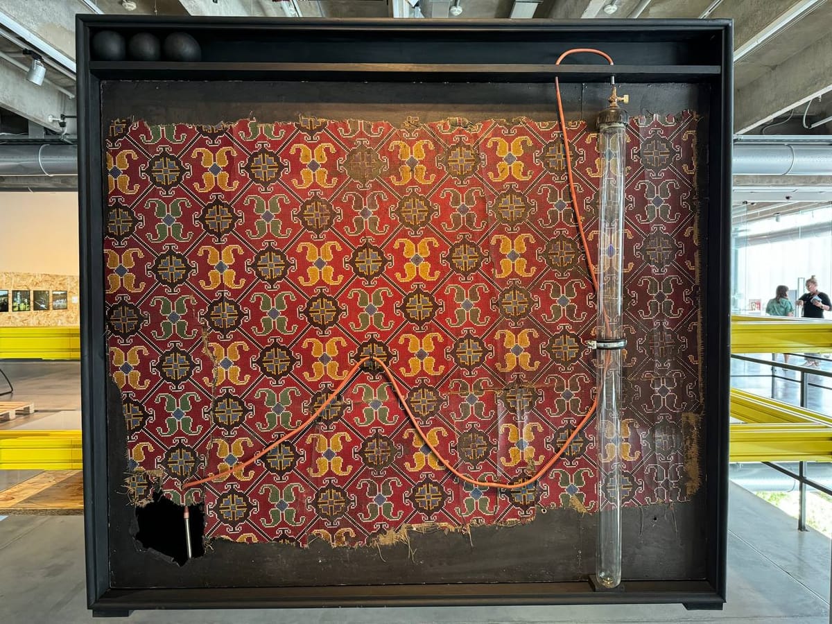 В чёрной витрине закреплён потёртый красный textile с вышитым крестиком орнаментом; рядом вертикальная стеклянная трубка, от неё тянется оранжевый шланг. В зале виден экспозиционный интерьер