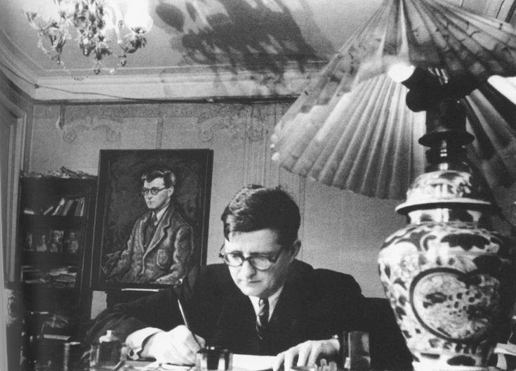 Мужчина в очках и тёмном костюме пишет за столом, на стене позади него портрет, справа лампа с плиссированным абажуром и расписной вазой.