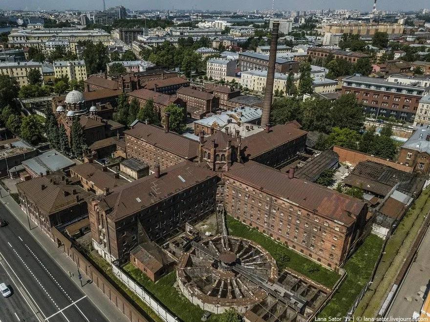 Красные кирпичные корпуса тюрьмы с высокой трубой, церковью с куполом и круглой постройкой без крыши; вокруг городская застройка и дорога.