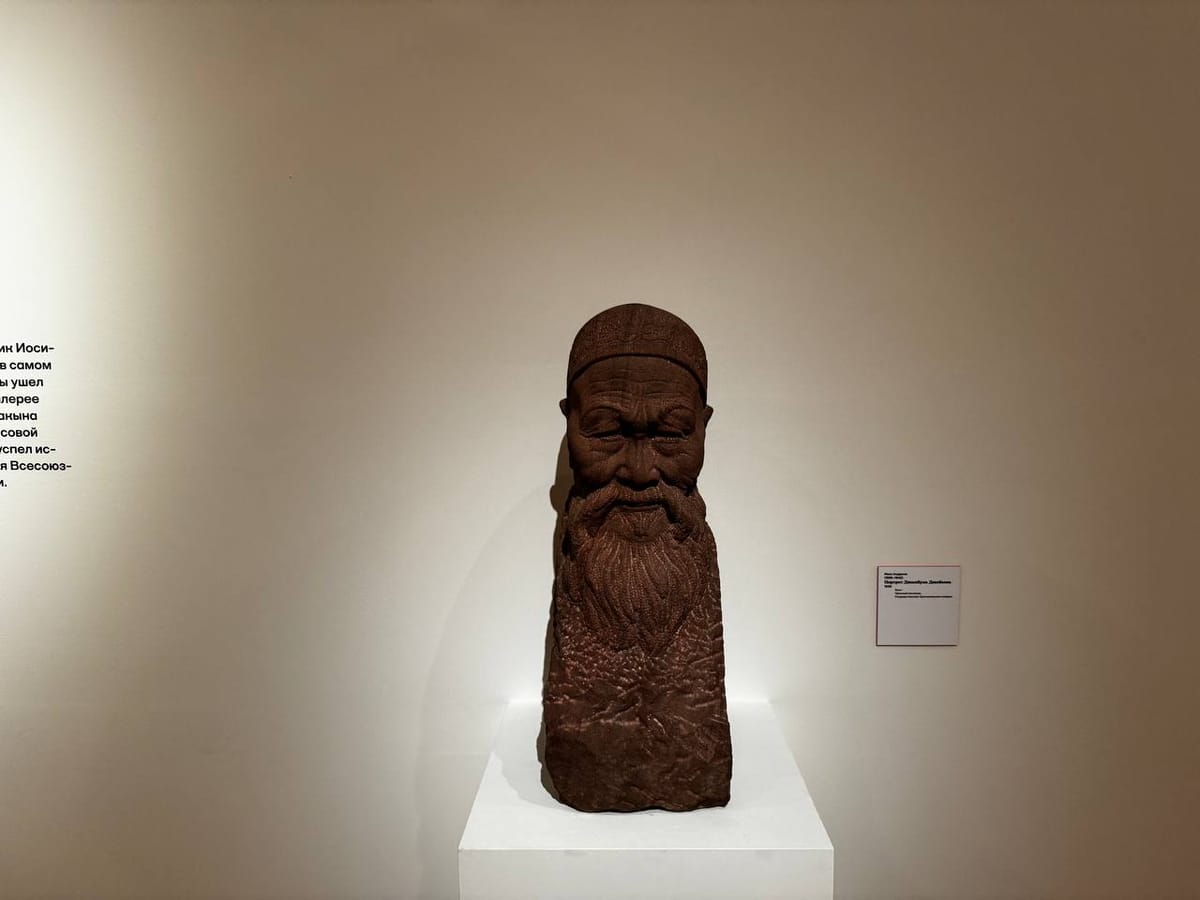 Скульптурный портрет старика с длинной бородой в тюбетейке из красновато-коричневого камня на белом постаменте у светлой стены музея. Справа висит табличка.