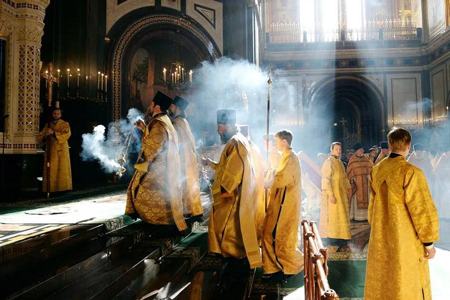 Священнослужители в золотых облачениях во время богослужения в соборе, лучи света пронизывают клубы кадильного дыма.