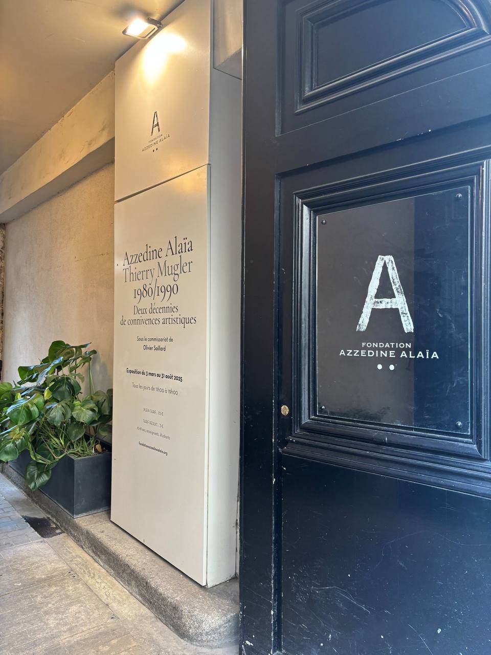 Чёрная дверь с логотипом Fondation Azzedine Alaïa и стенд с анонсом выставки «Аззедин Алайя, Тьерри Мюглер 1980/1990» (3 марта – 31 августа 2025). Рядом растение в кашпо.