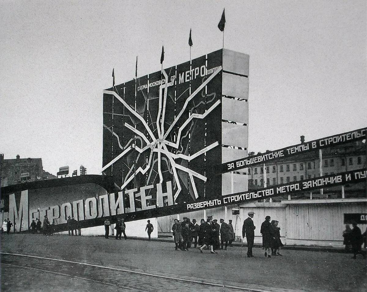 Огромный щит со схемой линий метро, надпись «Метрополитен» и ленты с лозунгами о темпах строительства. Перед ним идут прохожие, рядом дощатое ограждение стройки.