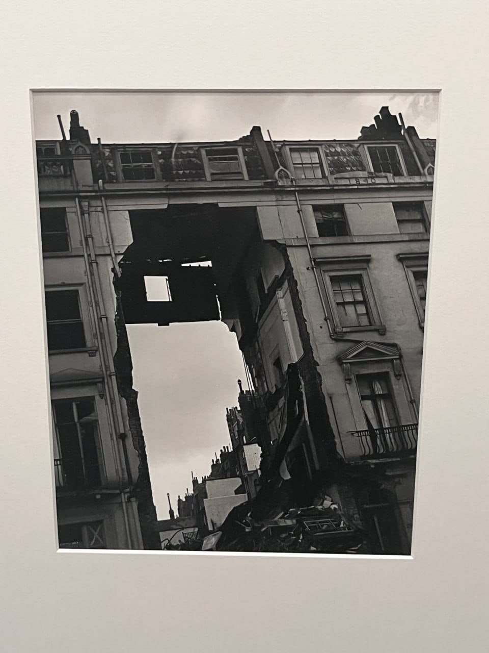 Чёрно-белая фотография на выставке: многоэтажный дом с выбитыми окнами, посреди фасада зияет провал до неба, внизу груда обломков.