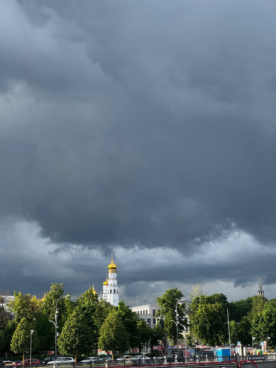 Тёмные грозовые тучи нависли над белой колокольней с золотым куполом. Внизу видны зелёные деревья, набережная с машинами и шпиль кремлёвской башни справа.