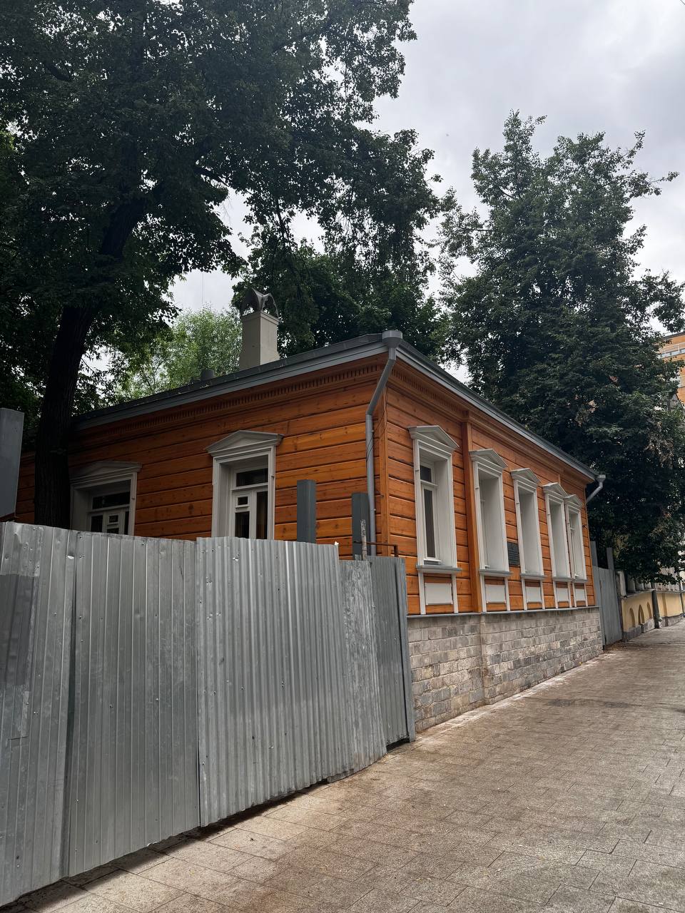 Одноэтажный деревянный дом оранжево-коричневого цвета с белыми наличниками и каменным цоколем. Он стоит на тихой улице под высокими деревьями, перед ним забор из профнастила.