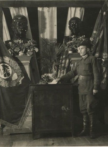 Старая чёрно-белая фотография: военный в форме и сапогах стоит у тумбы с цветами. Позади на постаментах два бюста с цветами, по сторонам тёмные знамёна.