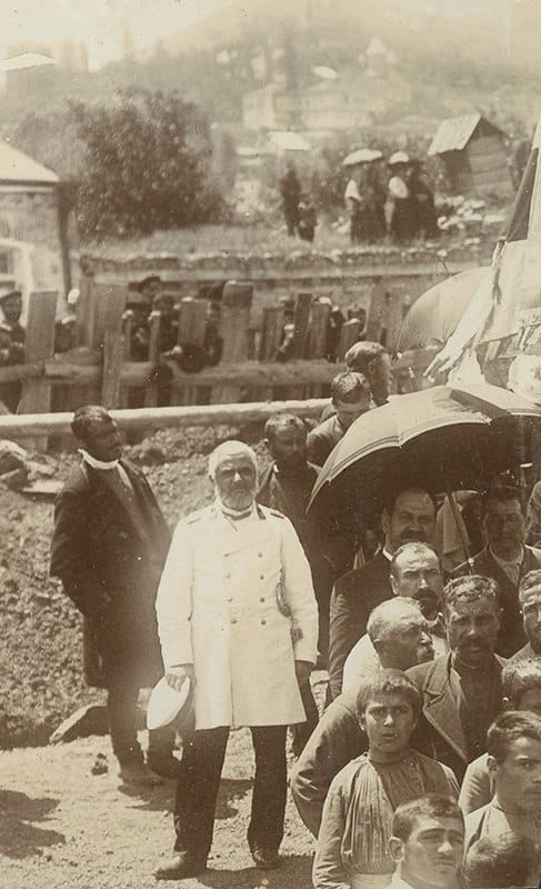 Старая фотография: пожилой бородатый мужчина в белом кителе стоит перед толпой мужчин и мальчиков, справа зонт и флаг, позади деревянная ограда и холм с домами.