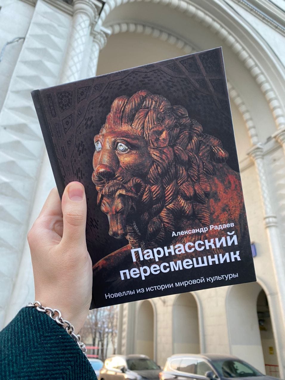 Рука держит книгу «Парнасский пересмешник» Александра Радаева с изображением львиной головы на фоне белого здания с аркой.