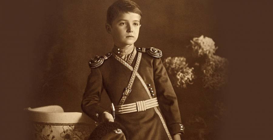 Сепия: мальчик в тёмной форме с эполетами, галунной перевязью и поясом стоит вполоборота, опираясь на вазу; рядом цветы хризантемы, в руке папаха.