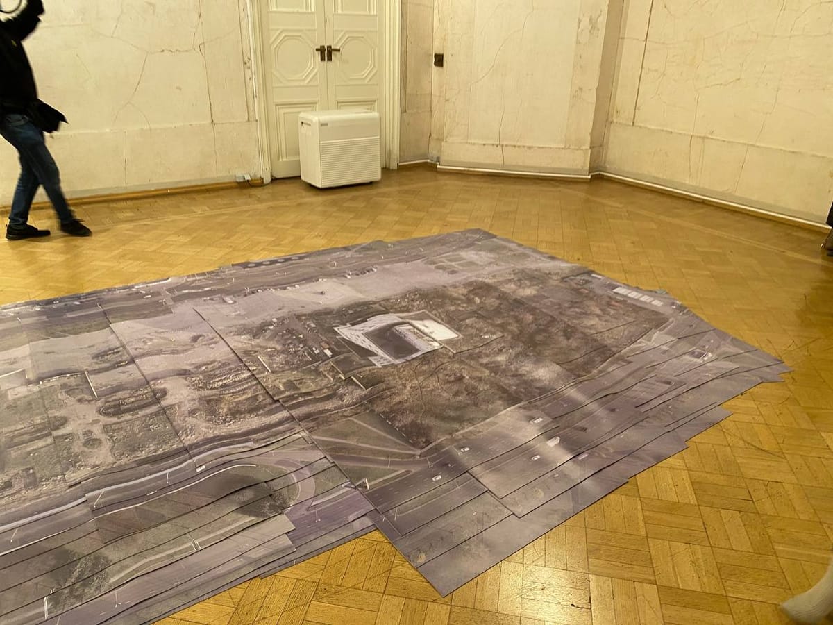 На паркетном полу старинного зала лежит наклеенная стопкой крупная аэрофотосъёмка с дорогой и зданием в центре. Слева идёт человек, у стены стоит белый прибор.