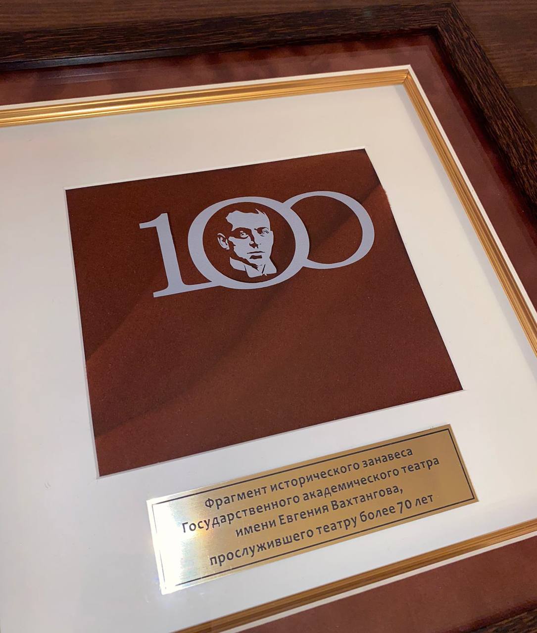 Рамка с фрагментом бордового занавеса, эмблемой «100» с портретом и золотистой табличкой о занавесе театра имени Вахтангова.