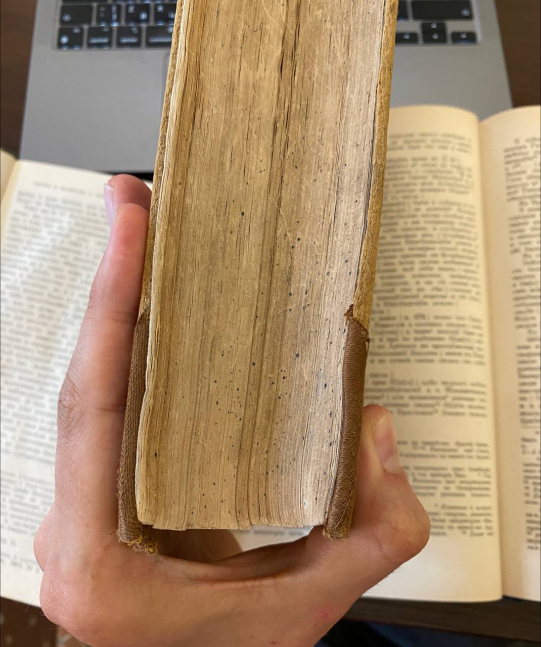 Рука держит толстую старую книгу с потёртым кожаным корешком, на заднем плане раскрытые книги и ноутбук.