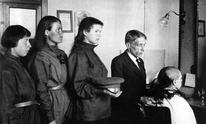 Чёрно-белый снимок: мужчина в костюме стрижет сидящую женщину, рядом в ряд стоят три женщины в гимнастёрках, одна держит фуражку.