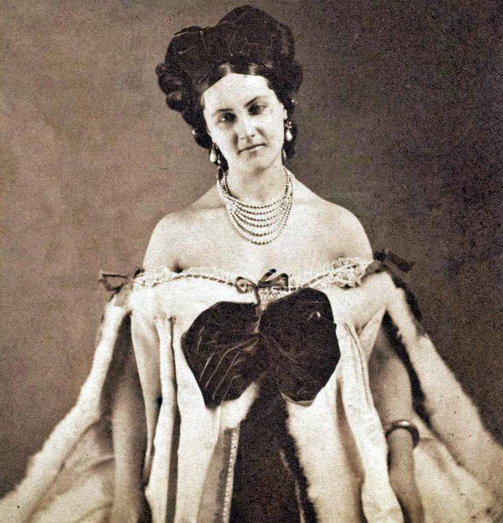 Старинная сепийная фотография: молодая женщина с высокой причёской, в серьгах и многорядном жемчужном ожерелье, в платье с открытыми плечами, меховой отделкой и большим тёмным бантом.