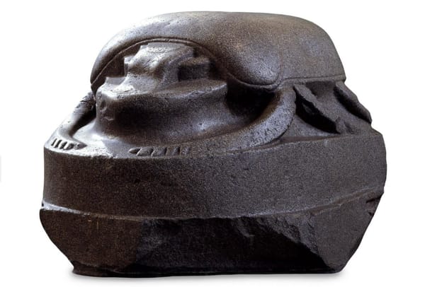 Крупная скульптура жука-скарабея из тёмного зернистого камня: гладкие надкрылья, согнутые лапки по бокам, грубо обработанное основание.