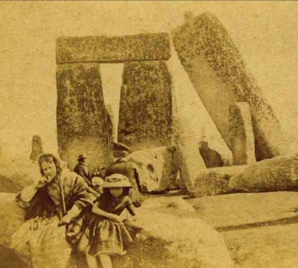 Сепия 1865 года: женщина и девочка в шляпке сидят перед громадными камнями Стоунхенджа, позади стоит мужчина в шляпе.
