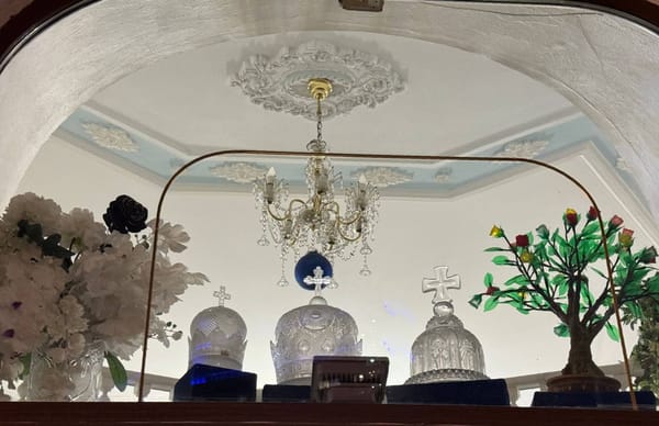 В витрине под лепным потолком с хрустальной люстрой стоят три хрустальные короны с крестами, слева белые цветы, справа дерево с яркими цветами.