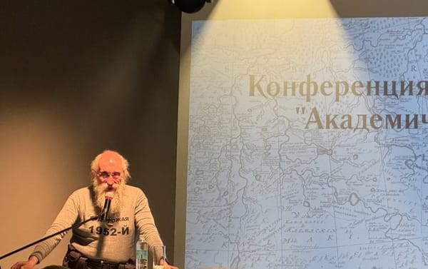 Седобородый мужчина в очках и серой кофте говорит в микрофон на фоне экрана со словом «Конференция» и старинной картой.