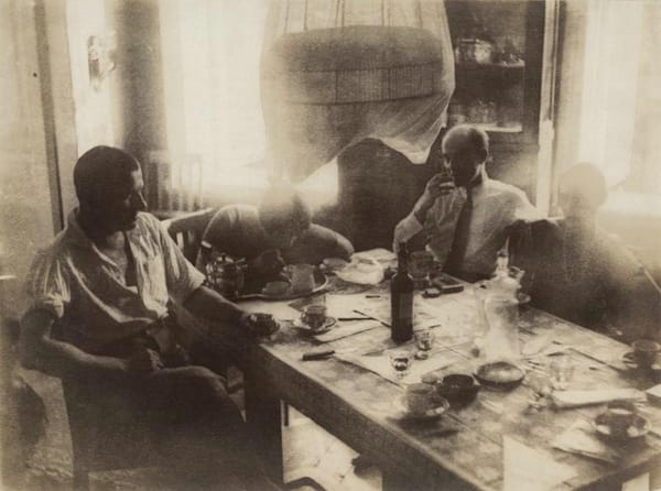 Старая сепийная фотография: несколько мужчин сидят за столом с чашками, бутылкой и графином, сверху виден полупрозрачный абажур.