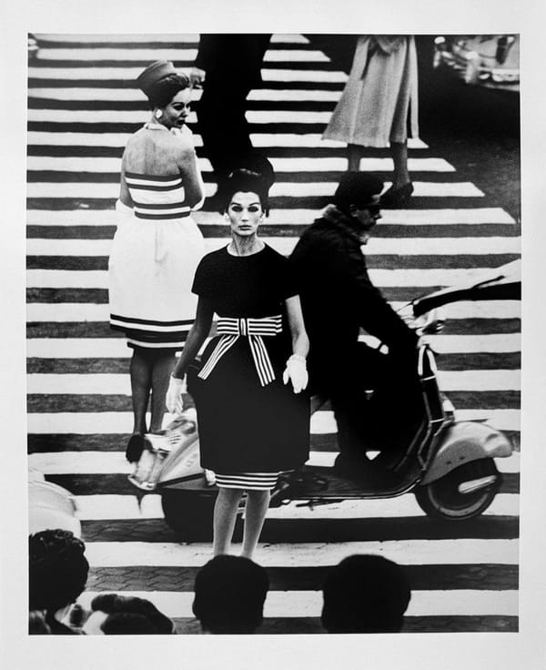 Чёрно-белый снимок: две женщины в платьях с полосатыми деталями стоят на зебре, мимо проезжает мужчина на веспе. Внизу кадра видны затылки прохожих.