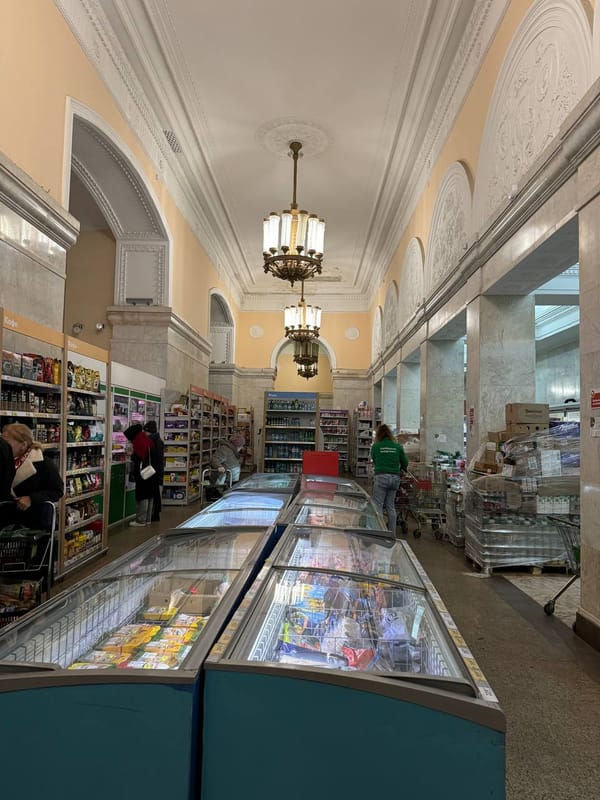Зал магазина с лепным потолком, люстрами, арками, морозильными витринами и покупателями между полками с товарами.