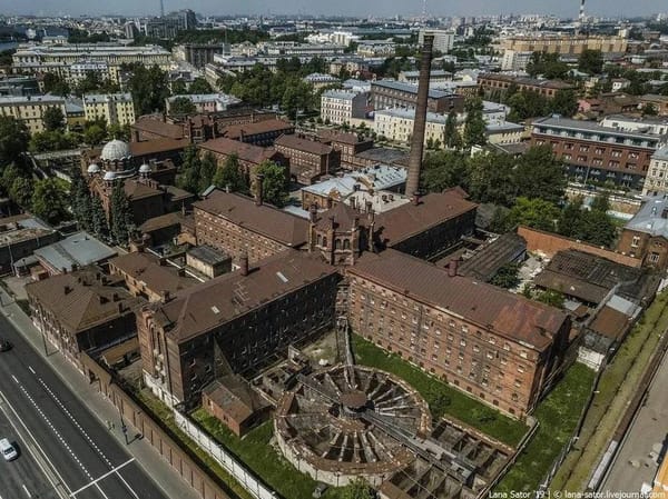Красные кирпичные корпуса тюрьмы с высокой трубой, церковью с куполом и круглой постройкой без крыши; вокруг городская застройка и дорога.
