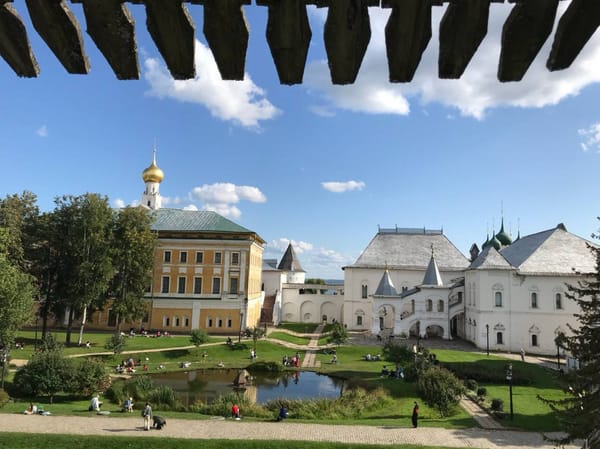 Двор Ростовского кремля с жёлтым административным зданием, белыми палатами, зелёными куполами, прудом и людьми на газонах.